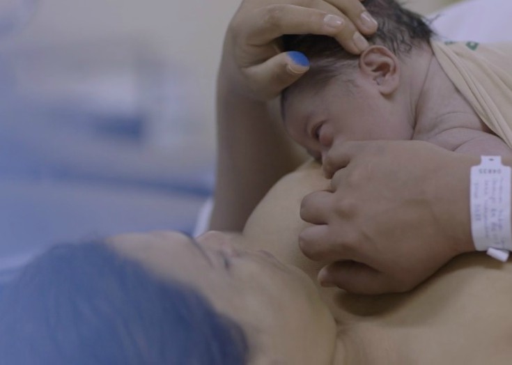 Uma mulher de cabelos castanhos deitada, com um recém nascido em seu colo. Na mão direita é possível ver uma pulseira de identificação do hospital.