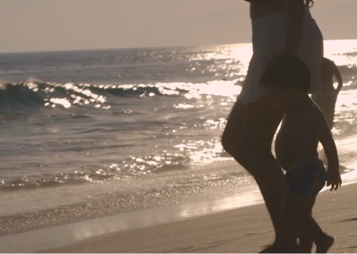 Uma criança segurando a mão da mãe, enquanto corre para o mar em uma praia.